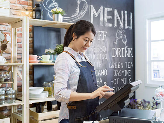 Female barista in apron using point of sale terminal to run her successful coffee store business